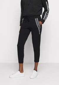 Černé zkrácené tepláky s páskem na stažení v pase, které mají bílé boční pruhy a detaily loga, vyrobené z měkké, elastické tkaniny.