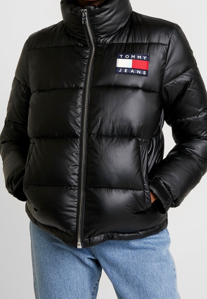 Zwarte pufferjack met hoge kraag, ritssluiting aan de voorkant en horizontale quilting. Voorzien van een logopatch met rode, witte en blauwe accenten.