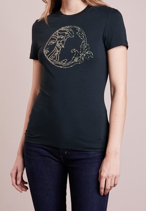 Femme portant un t-shirt noir ajusté avec un motif circulaire de vagues dorées à clous sur la poitrine, associé à un jean bleu foncé.
