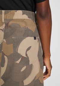 Camo-patterned pants in beige and olive, made of sturdy fabric, featuring a back pocket and a branded label near the waistband.