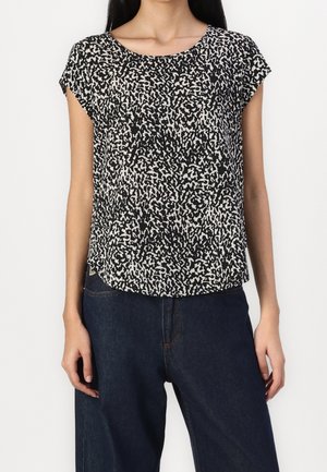 Femme portant un chemisier à manches courtes à motif abstrait noir et blanc, associé à un jean taille haute bleu foncé, debout devant un fond blanc.