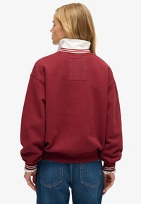 Mujer rubia con sudadera roja de cuello y puños a rayas blancas, vistiendo jeans azules, de espaldas contra un fondo blanco liso.