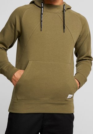 Homme portant un sweat à capuche à enfiler couleur vert olive avec des cordons noirs et une poche kangourou à l'avant, les mains dans les poches.