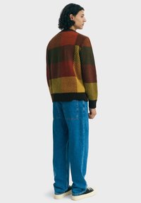 Cardigan en tricot avec un grand motif à carreaux multicolore en rouge, jaune, vert et marron. Manches à côtes et ourlet, associé à un pantalon en denim bleu.