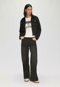 Gestructureerde zwarte cropped jas met drukknoopjes, witte graphic t-shirt, donkere wijde broek en beige schoenen met rubberen zolen.