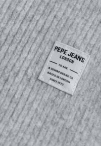 Etiqueta de tela blanca con texto negro que dice "PEPE JEANS LONDON", cosida sobre un fondo gris texturizado, detallando el origen y la historia de la marca.