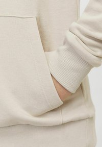 Felpa beige con tasca frontale, polsini a coste e tessuto testurizzato. Presenta una vestibilità rilassata e dettagli delle cuciture minimi.