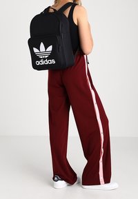 Black Adidas backpack with white logo, worn on the back. Red wide-leg pants with pink buttons along the side. White sneakers.