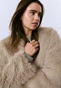 Joven mujer con cabello largo y castaño, vestida con un abrigo de piel sintética beige, mirando de lado con la mano cerca del cuello.