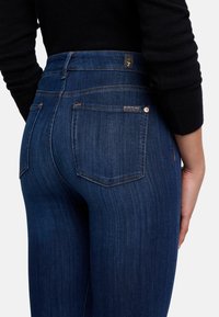 Jean en denim bleu foncé à taille haute, avec deux poches arrière et de subtils détails de couture. Texture lisse, design ajusté.