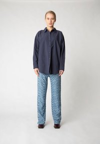 Marineblaue Hemd mit Knöpfen in lockerer Passform, kombiniert mit gemusterten hellblauen Hosen, die ein strukturiertes Design aufweisen, und braunen Schuhen.