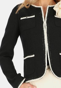 Czarna kurtka z białymi wykończeniami, teksturowany materiał, dwie przednie kieszenie i dopasowany krój. Posiada okrągły dekolt oraz widoczne detale szwów.