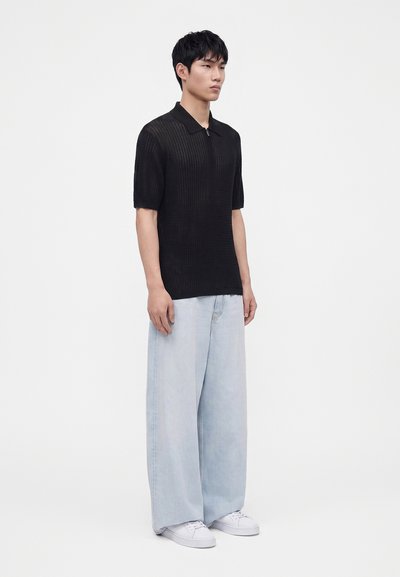 Jeune homme debout portant un t-shirt noir à manches courtes zippé, un jean large bleu clair et des baskets blanches sur un fond uni clair.