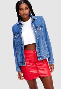 Femme aux longs cheveux tressés portant une veste en jean bleue, un t-shirt blanc court et une mini-jupe en cuir rouge vif avec une fermeture éclair à l'avant.