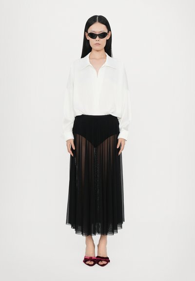 Blusa blanca con escote en V y corte holgado, combinada con una falda plisada negra transparente y sandalias de tacón de terciopelo burdeos. Gafas de sol negras incluidas.