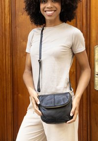 Musta nahkainen olkalaukku vetoketjulla, jossa on pyöreä muoto ja irrotettava olkahihna. Kuvioitu pinta lisää visuaalista mielenkiintoa.