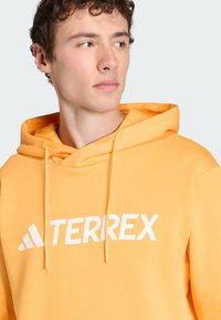 Pomarańczowa bluza z kapturem wykonana z miękkiego materiału, z kieszenią kangurzą, kapturem ze sznurkiem i dużym białym logo "TERREX" z przodu.