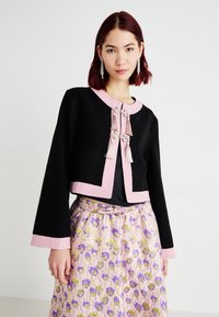 Jachetă neagră scurtă cu dantelă roz, având închideri decorative, țesătură texturată și mâneci lungi. Pură peste o fustă cu model.