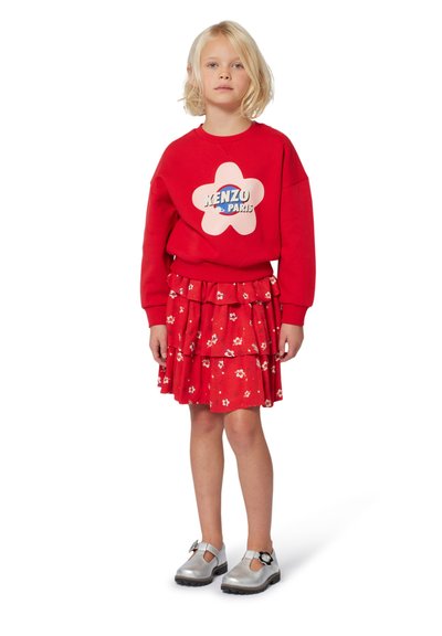 Felpa rossa con logo a fiore rosa, gonna rossa a strati con motivo floreale e scarpe argentate con dettaglio a fibbia.