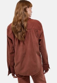 Chemise en velours côtelé couleur rouille avec une coupe ample, poignets à boutons et un dos plissé. Texture lisse et détail d'ourlet arrondi.