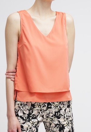 Bezrekawnik w kolorze koralowym z dekoltem w kształcie litery V połączony z czarno-białymi spodniami w kwiatowy wzór na osobie na jednolitym tle.