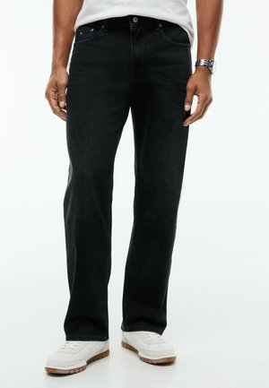 Mand iført sorte jeans med lige ben, hvide sneakers, hvid indstoppet skjorte og et sølvur på venstre håndled, stående mod en hvid baggrund.