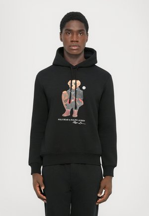 Joven con sudadera negra con un gráfico de un oso en un atuendo de invierno sosteniendo unas raquetas de nieve y el texto "Polo Bear de Ralph Lauren."