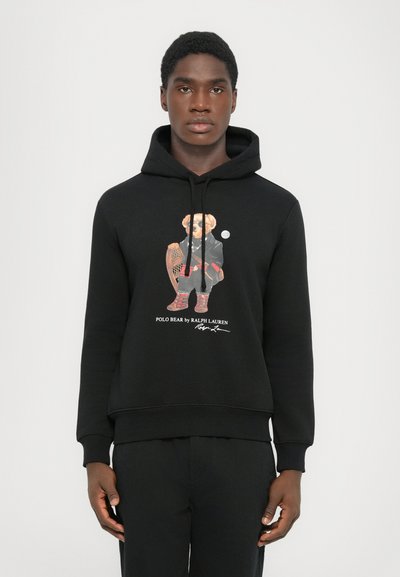 Joven con sudadera negra con un gráfico de un oso en un atuendo de invierno sosteniendo unas raquetas de nieve y el texto "Polo Bear de Ralph Lauren."