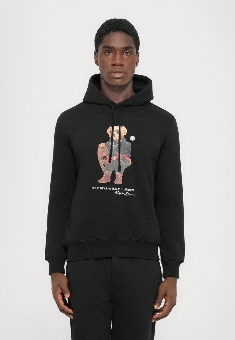 Mladý muž v černé mikině s kapucí, na které je grafika medvěda v zimním oblečení, jak drží sněžnice, a nápis "Polo Bear by Ralph Lauren."