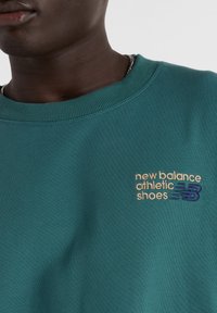 Sudadera verde con cuello de canalé, textura suave y logotipo bordado de "New Balance Athletic Shoes" en dorado y azul en el pecho.