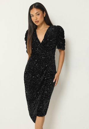 Donna che indossa un vestito nero aderente con paillettes, maniche corte a sbuffo e scollo a V, in piedi davanti a uno sfondo chiaro e semplice.