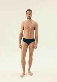 Czarne kąpielówki z gładkiego materiału, dopasowany krój z niskim stanem i minimalnym logo z boku. Model stoi boso.