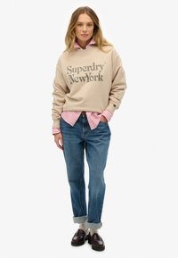 Mujer vestida con un sudadera beige de Superdry de Nueva York sobre una camisa de rayas rosas, jeans azules remangados, calcetines blancos y mocasines marrones oscuros, de pie con la mano en el bolsillo.
