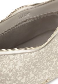 Bolso de textura beige con un patrón en espiral, cierre de cremallera y forro interno de tela. Presenta el logotipo "TOUS" en relieve y acentos de hardware en tono dorado.
