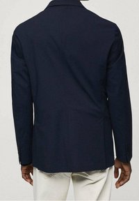 Granatowa marynarka z teksturowanej tkaniny, z pojedynczym tylnym rozventylowaniem, klapami z ząbkami i mankietami na guziki, zestawiona z jasnymi spodniami.