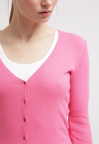 Pink knitted cardigan with a V-neck, featuring a row of buttons down the front and long sleeves, worn over a white top.
