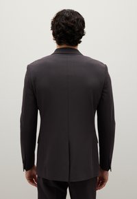 Giacca da abito grigio scuro, confezionata su misura, con una superficie liscia, spalle strutturate e senza motivi visibili, caratterizzata da un unico spacco sul retro.