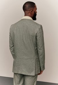 Abito verde chiaro su misura con tessuto strutturato. Presenta un colletto color crema e due spacco posteriori. Vestibilità slim con maniche lunghe.