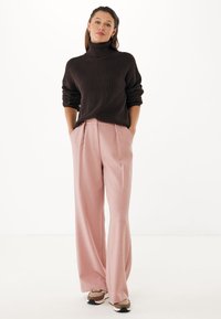 Suéter marrón de cuello alto acanalado combinado con pantalones de pierna ancha rosa claro, con plisados frontales y bolsillos laterales, llevados con zapatillas beige y blancas.