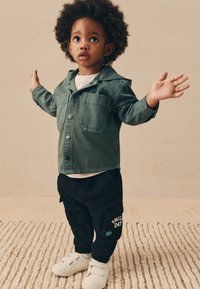 Jachetă verde din denim cu două buzunare frontale, pantaloni cargo negri cu detaliu text alb și adidași albi. Textură plată pe un covor țesut.