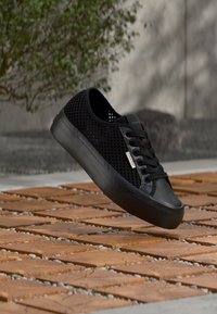 Μαύρο αθλητικό παπούτσι Calvin Klein με πλευρές από δίχτυ και κορδόνια που αιωρείται πάνω από ξύλινα πλακάκια και χαλίκι σε εξωτερικό χώρο δίπλα σε δέντρο και τσιμεντένιο τοίχο.