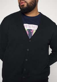 Svart cardigan med V-hals, knäppning fram och mjuk textur. Bärs över en marinblå t-shirt med grafik och färgglad design.