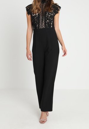 Kvinde iført en sort jumpsuit med blonder floral top og brede, ensfarvede bukser, sat sammen med nøgenfarvede åbne hæle, stående på en hvid baggrund.