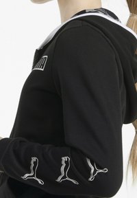 Mujer vestida con sudadera negra de Puma con logo blanco en el pecho y múltiples logos plateados del gato Puma en la manga, de pie de lado.