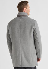 Grau melierter Wollmantel mit hohem Kragen, nahtlosem Design und seitlichen Taschen. Der Stoff hat eine glatte Textur mit dezenten Nahtdetails.