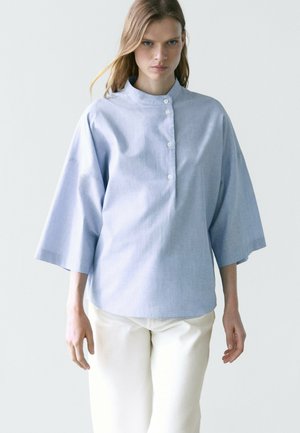 Femme portant une chemise ample bleu clair à boutons avec des manches trois quarts et un pantalon de couleur crème, debout devant un fond uni.