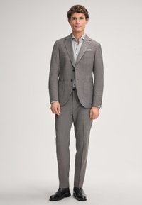 Mężczyzna ubrany w dopasowany szary garnitur, jasnoszary koszuli i czarne eleganckie buty, stojący na tle jasnego, jednolitego tła.