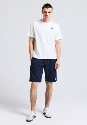 Giovane uomo in piedi che indossa una maglietta bianca a maniche corte, pantaloncini blu scuro e scarpe da ginnastica bianche con strisce verdi su uno sfondo chiaro uniforme.