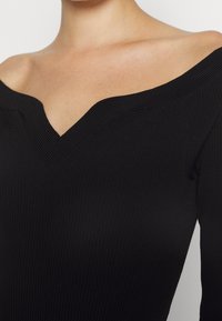 Femme portant un haut noir côtelé à épaules dénudées avec un décolleté en V, laissant apparaître la peau nue sur le haut de la poitrine et les épaules.