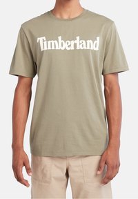 Lys olivengrøn bomulds t-shirt med korte ærmer og rund hals, prydet med "Timberland" trykt i fede hvide bogstaver over brystet.